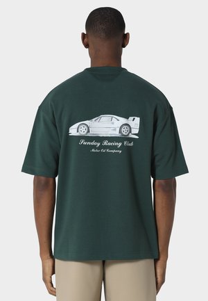 Homme portant un T-shirt vert foncé avec une voiture de sport blanche et le texte « Sunday Racing Club Motor Oil Company » dans le dos, pantalon beige.