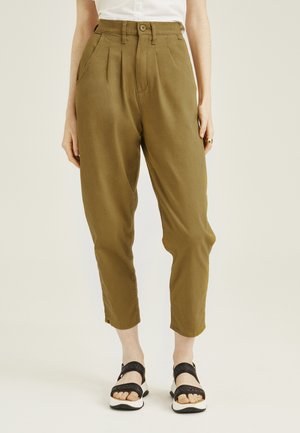 Femme portant un pantalon fuselé taille haute vert olive avec des plis, des poches latérales, une fermeture par bouton et zip, associé à des sandales noires.
