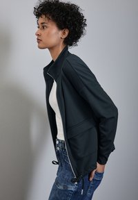 Giacca nera con zip e colletto alto, tessuto leggero, tasche laterali e orlo con coulisse, indossata sopra una maglietta bianca e jeans blu.