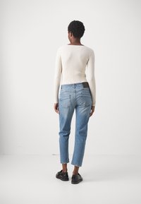 Lichtbeige geribde langemouwen top en vervaagde blauwe spijkerbroek met een rechte snit. Zwarte schoenen maken de outfit af. Achteraanzicht tegen een witte achtergrond.