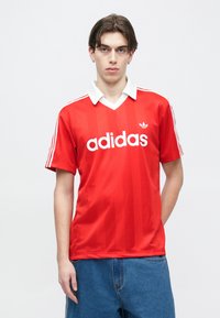 Νεαρός άνδρας με μεσαίου μήκους σκούρα μαλλιά, φορώντας κόκκινο μπλουζάκι Adidas με λευκό γιακά και ρίγες, και μπλε τζιν, στέκεται μπροστά σε απλό φόντο.