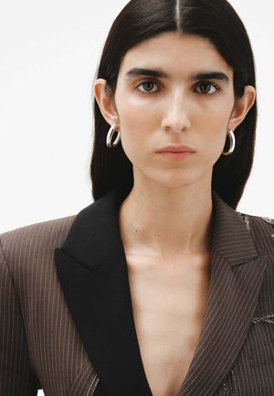 Blazer marrón de rayas con solapa negra. La modelo lleva pendientes grandes de aro plateados. Textura suave, detalle de costura visible en el cuello.