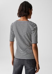 Femme aux cheveux courts et foncés portant une chemise à manches courtes noir et blanc à rayures et un jean gris foncé, vue de dos sur un fond uni.