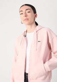 Hoodie zippé rose clair en tissu texturé, avec des poches avant et un petit logo vert sur la poitrine. Porté sur une chemise blanche.