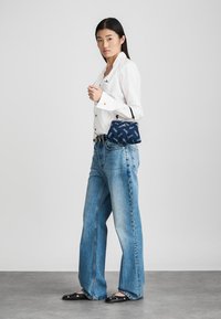 Mulher vestindo uma camisa branca de botão e jeans largos azuis, segurando uma pequena bolsa marinha com um padrão, em pé sobre um chão cinza.