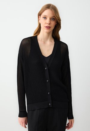 Donna con capelli castano chiaro che indossa un cardigan nero strutturato con bottoni sopra una maglietta nera, in piedi contro uno sfondo leggero e uniforme.