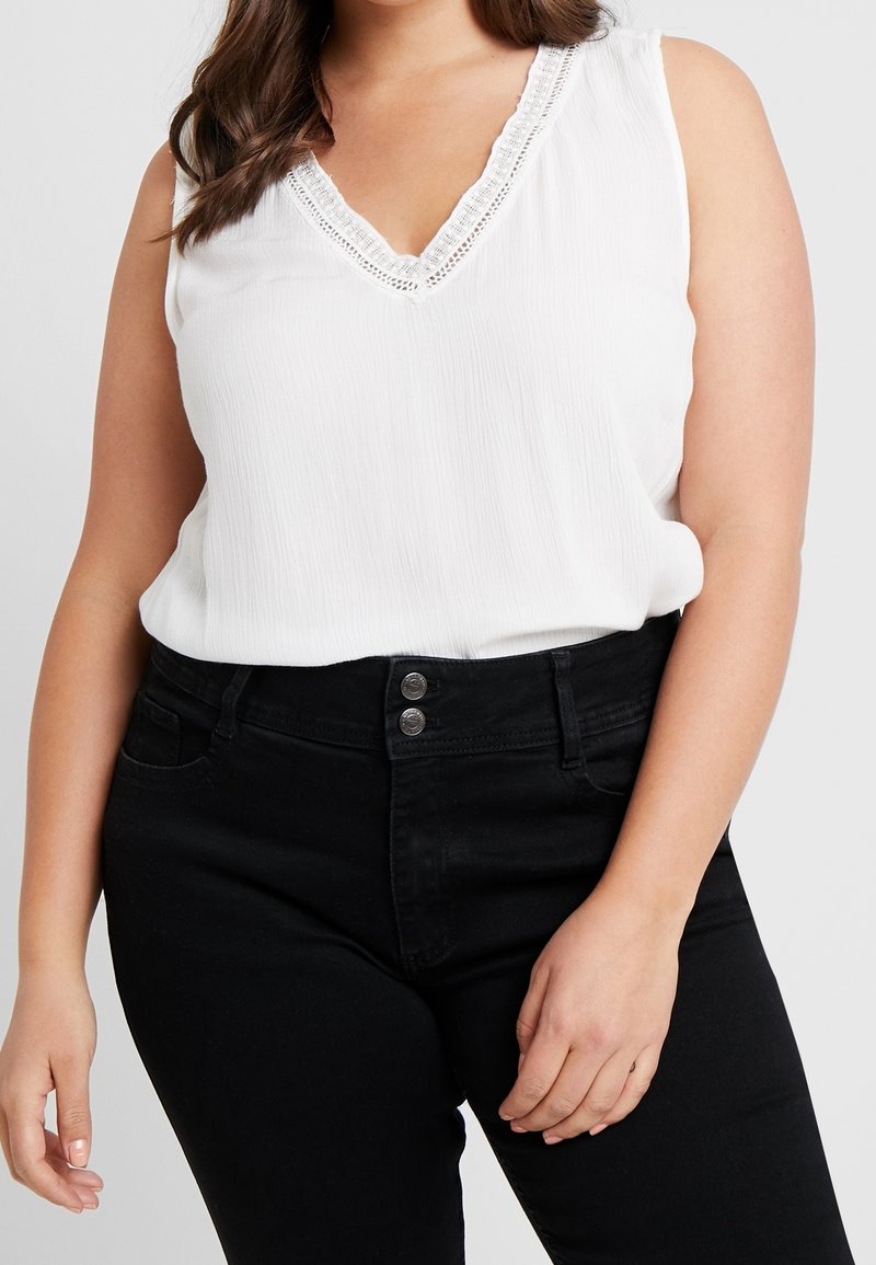 Top bianco senza maniche con scollatura a V e bordo in pizzo, abbinato a jeans neri a vita alta con chiusura a due bottini argentati.
