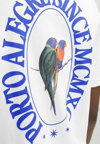 Camiseta blanca con una colorida ilustración de dos loros posados en una rama, rodeados de texto circular azul.