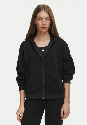 Mujer con pelo largo y liso que lleva una sudadera con capucha negra con cremallera y pantalones negros sobre un fondo claro y liso.
