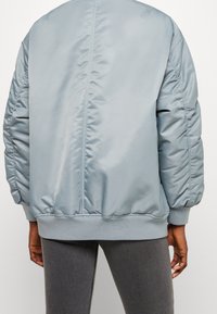 Giacca bomber azzurro chiaro con texture trapuntata, polsini e orlo a coste. La vista posteriore mostra un tessuto liscio e dettagli delle cuciture.