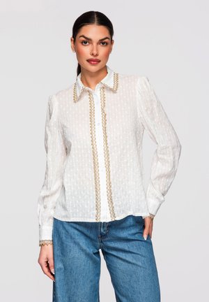 Vrouw met donker haar in een staart, gekleed in een witte blouse met textuur en gouden randje, en een blauwe spijkerbroek, staand tegen een effen lichte achtergrond.