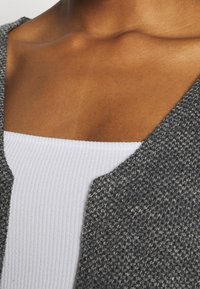 Šedý texturovaný cardigan s výstřihem, který je přes bílý ribovaný top, ukazuje strukturu tkaniny a hladké kontury.