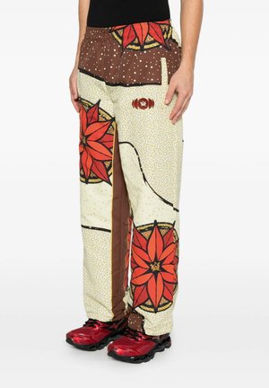 Persona che indossa pantaloni fantasia con grandi disegni floreali rossi e sezioni marroni, beige e gialle abbinati a scarpe sportive rosse e nere.
