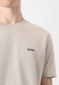 Beige bavlněné tričko s žebrovaným kulatým výstřihem a malým černým logem "BOSS" na levé části hrudi. Hladká textura, krátké rukávy.