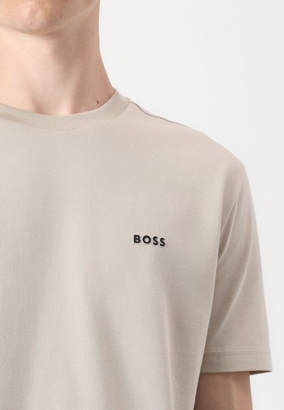 T-shirt en coton beige avec un col rond côtelé et un petit logo noir "BOSS" sur la poitrine gauche. Texture lisse, manches courtes.