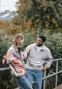 Dwie osoby na moście: jedna w wielokolorowej kurtce puchowej, a druga w szarej polarowej bluzie, obie w dżinsach, otoczone zielenią.