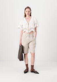 Biele skrátené blúzky s vyrezávanými detailmi, béžové klasické šortky, hnedé členkové ponožky a čierne baleríny. Modelka drží hnedú kabelku.