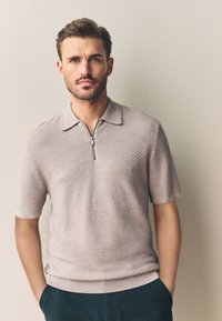 Polo à manches courtes texturé de couleur beige clair avec un col zippé. Tissu doux et ourlet côtelé, porté avec un pantalon foncé.