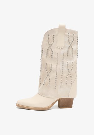 Botín de ante beige con punta afilada, tachuelas plateadas decorativas en un patrón y un tacón de bloque de madera. Presenta una lengüeta trasera.