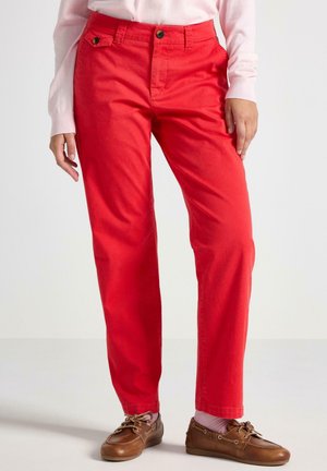 Donna con pantaloni rossi vivaci a gamba dritta, un maglione rosa e scarpe da barca marroni, in piedi contro uno sfondo bianco.