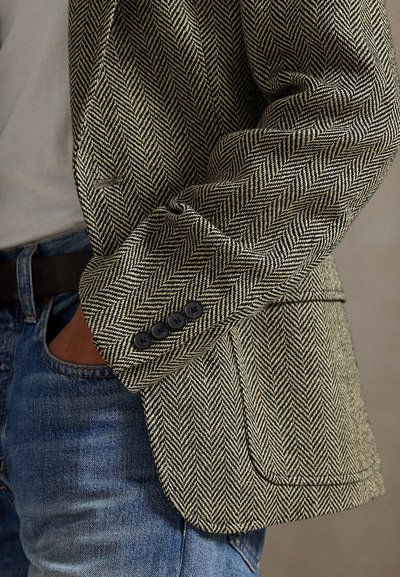 Persoon draagt een bruin en zwart blazer met visgraatpatroon en zwarte knopen, blauwe spijkerbroek en een beige overhemd, hand in de zak.