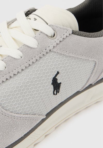 Ανοιχτό γκρι αθλητικό παπούτσι με λευκά κορδόνια, δίχτυ στο πλευρικό πάνελ, σουέντ λεπτομέρειες και μαύρο λογότυπο παίκτη πόλο στην εξωτερική πλευρά.