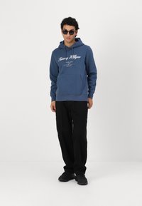 Hanorac albastru din bumbac, cu o buzunar frontal și un logo alb. Asortat cu pantaloni negri și adidași negri, modelul poartă ochelari de soare. Fundal simplu.
