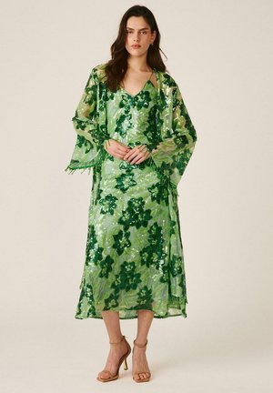 Mujer con vestido midi verde con lentejuelas florales y chal transparente a juego, de pie con las manos entrelazadas y sandalias de tacón alto color cuero claro.