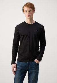 Sort langærmet t-shirt lavet af blødt materiale med rund halsudskæring og et lille broderet logo på brystet; sammen med blå jeans.