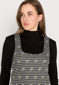 Vestido xadrez em preto e branco com padrão de flores roxas. Apresenta um decote quadrado e alças largas. Usado sobre um suéter de gola alta preto.
