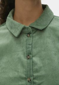 Detailní záběr osoby v světle zelené kordové košili s hnědými knoflíky a zakulaceným límcem.