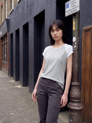 Jeune femme aux longs cheveux foncés portant un t-shirt gris et blanc et un pantalon foncé, debout, appuyée contre un lampadaire sur un trottoir en ville.