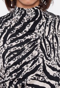 Blusa bianca e nera caratterizzata da un tessuto liscio con un audace motivo a strisce zebrate, colletto alto e dettagli arricciati sulle spalle.