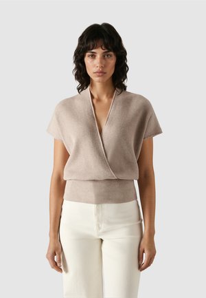 Frau mit dunklem, welligem Haar, die einen beigen Wickelpullover mit kurzen Ärmeln und hochtaillierte, cremeweiße Hose vor einem schlichten Hintergrund trägt.