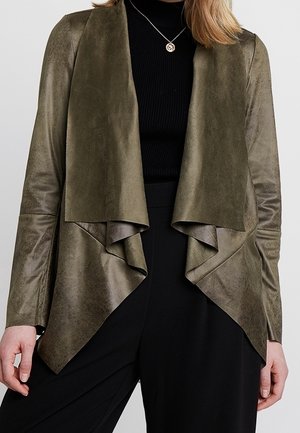 Woman wearing an olive green suede draped open-front jacket over a black turtleneck and black pants, with a pendant necklace.