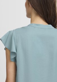 Blouse bleu clair avec des manches courtes à volants, un col rond et un tissu lisse légèrement texturé. Le dos est simple et sans ornement.