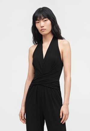 Femme aux longs cheveux noirs portant une combinaison noire sans manches avec un col licou et un détail plissé à la taille.