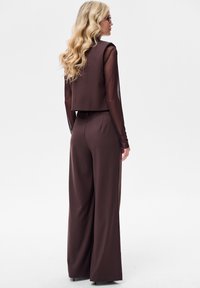 Brązowa krótka kurtka z przezroczystymi rękawami i pasujące szerokie spodnie. Gładka faktura, dopasowany krój. Noszona z długimi, falującymi włosami i okularami.