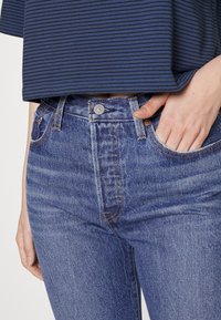 Jeans de mezclilla azul de cintura alta con costuras visibles, bolsillos delanteros y detalle de botón de metal. Combinados con una blusa de rayas en azul marino y gris.