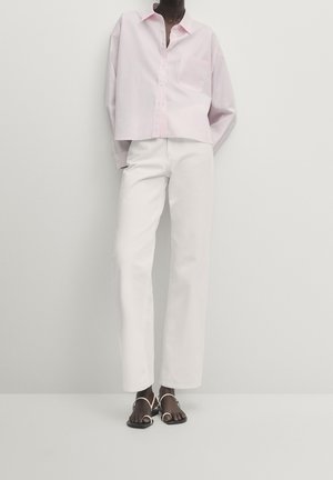 Person trägt ein locker sitzendes hellrosa Hemd mit Knöpfen, hochtaillierte gerade weiße Hose und weiße Sandalen mit Riemchen vor einfachem Hintergrund.