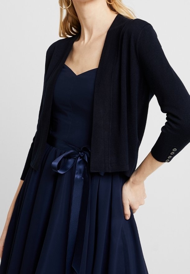 Femme portant une robe bleu marine avec une ceinture en satin nouée en nœud et un cardigan noir court avec trois boutons sur la manche.