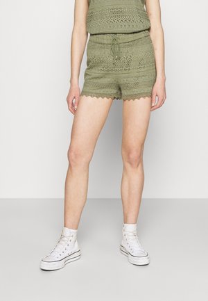 Kvinde iført olivengrøn hæklede shorts og matchende top med bølgekant, parret med hvide højtstående sneakers, stående på gråt gulv.