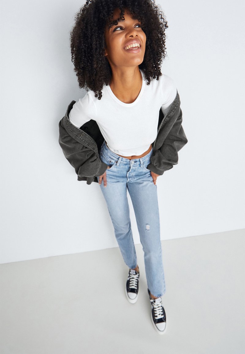 Top corto blanco, chaqueta verde oliva, jeans claros con detalle desgastado, zapatillas negras y blancas, tomado desde un ángulo alto.