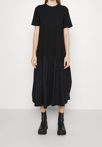 Schwarzes kurzärmeliges Kleid mit gerafftem unteren Saum, aus leichtem Stoff gefertigt. Getragen mit schwarzen klobigen Knöchelstiefeln.
