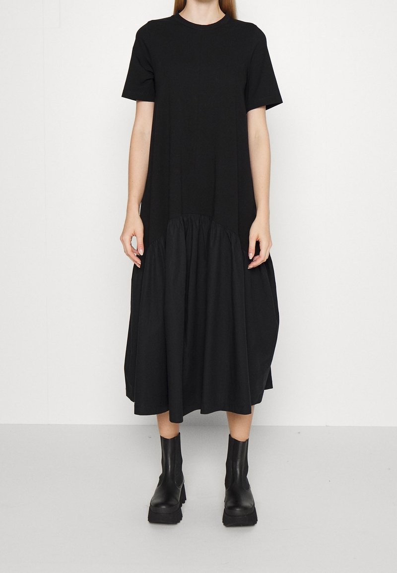 Schwarzes kurzärmeliges Kleid mit gerafftem unteren Saum, aus leichtem Stoff gefertigt. Getragen mit schwarzen klobigen Knöchelstiefeln.