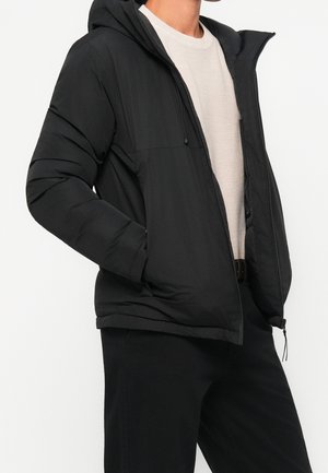 Personne portant une veste matelassée noire avec capuche, ouverte pour montrer une chemise beige texturée à manches longues en dessous.