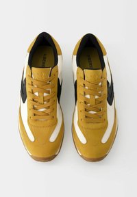 Gula och vita träningsskor med svarta detaljer, med rundad tå, snörning och en texturerad sula.