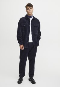 Navy corduroy shirt jacket with two chest pockets, button closure, and matching pants. Model wears a white t-shirt underneath and black shoes.