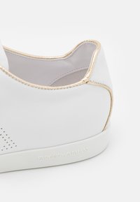 Sneaker in pelle bianca con accenti metallici oro lungo il collo e il tallone, caratterizzato da dettagli perforati e suola con marchio.