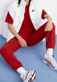 Roter Sweatshirt und passende Jogginghose, kombiniert mit einer weißen Steppweste. Die Schuhe haben ein graues und rotes Design mit mehrfarbigen Akzenten.